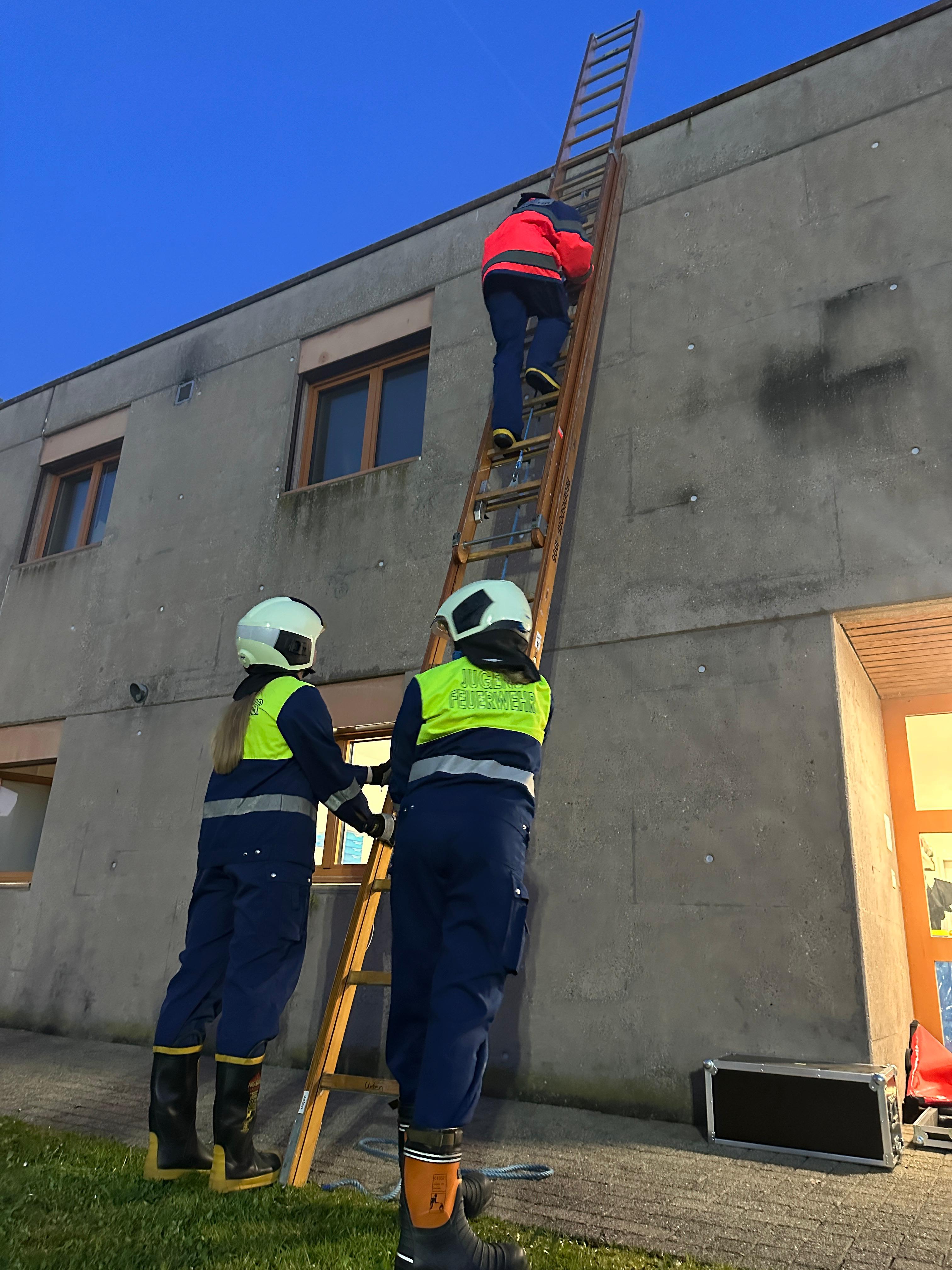 Zwei Jugendliche der Jugendfeuerwehr unteres Furttal sichern eine Leiter, während ein Kamerad zur Fassade aufsteigt – Abenddämmerung